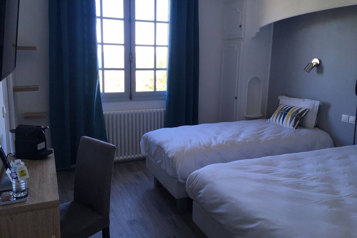 A hotel room with two beds, a desk with a chair, a radiator, and a window with curtains.