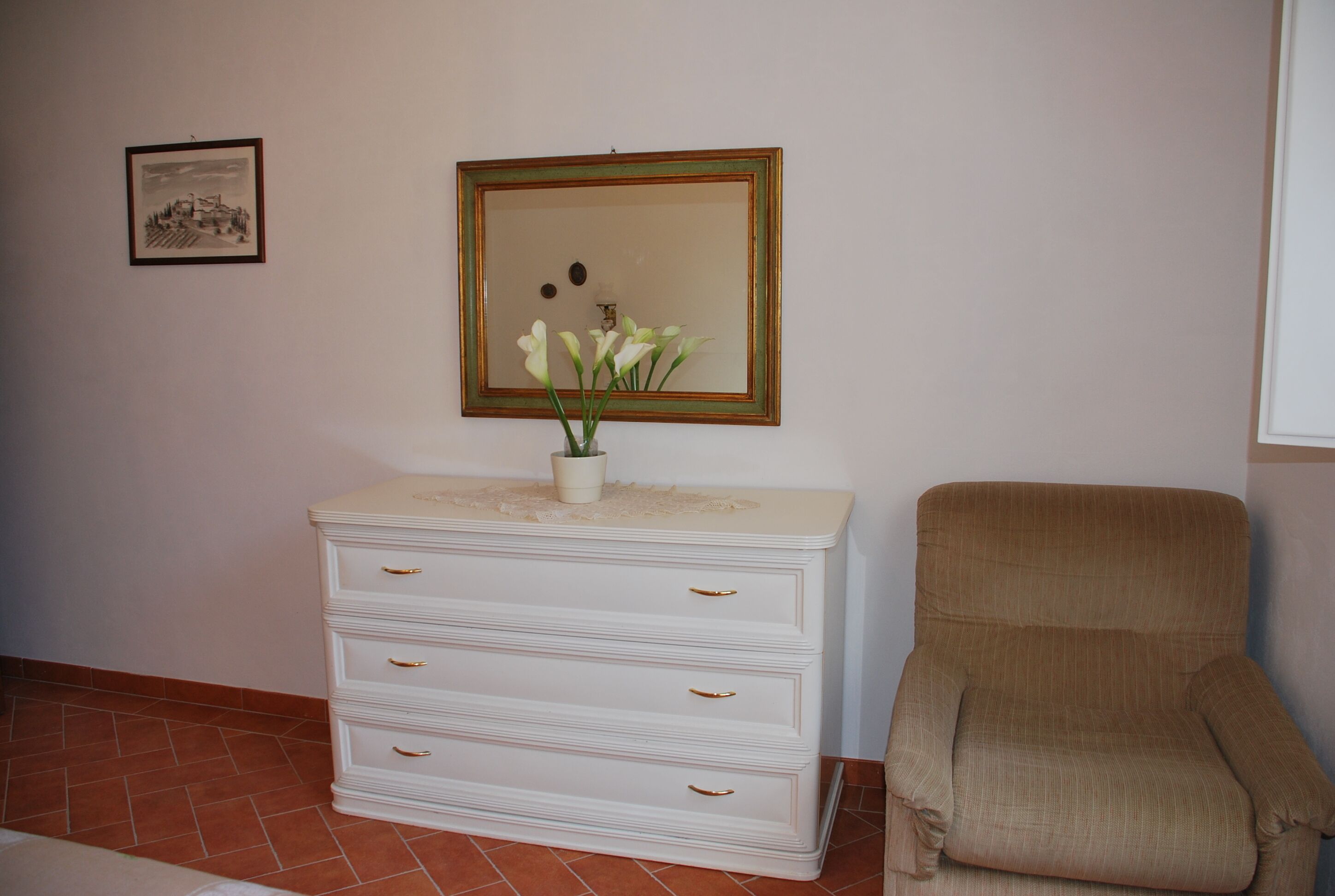 Une pièce avec une commode blanche, un tableau encadré au mur et un fauteuil beige.