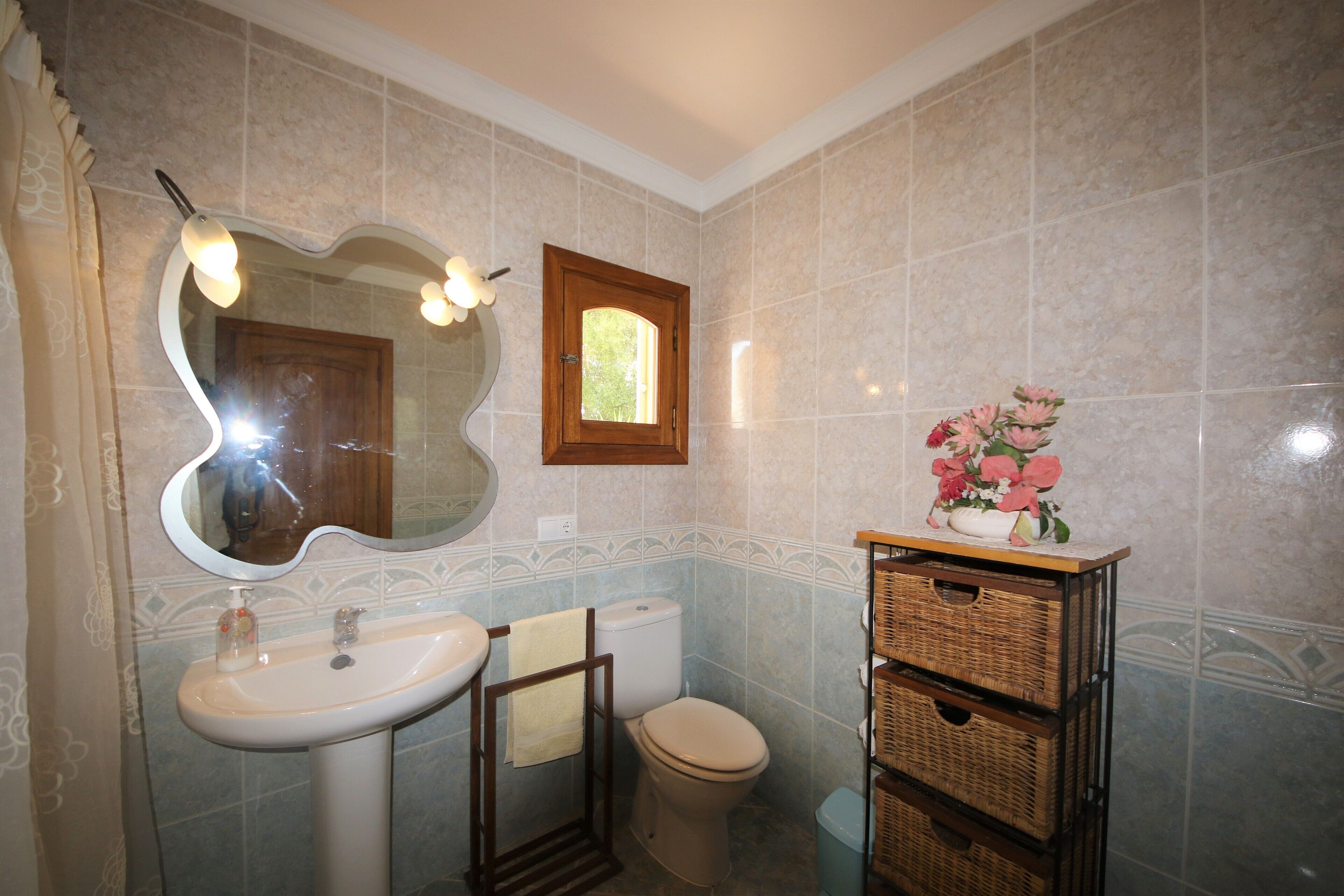 A bathroom with a sink, toilet, and a mirror with lights. There is a window and a vase with flowers on a shelf.