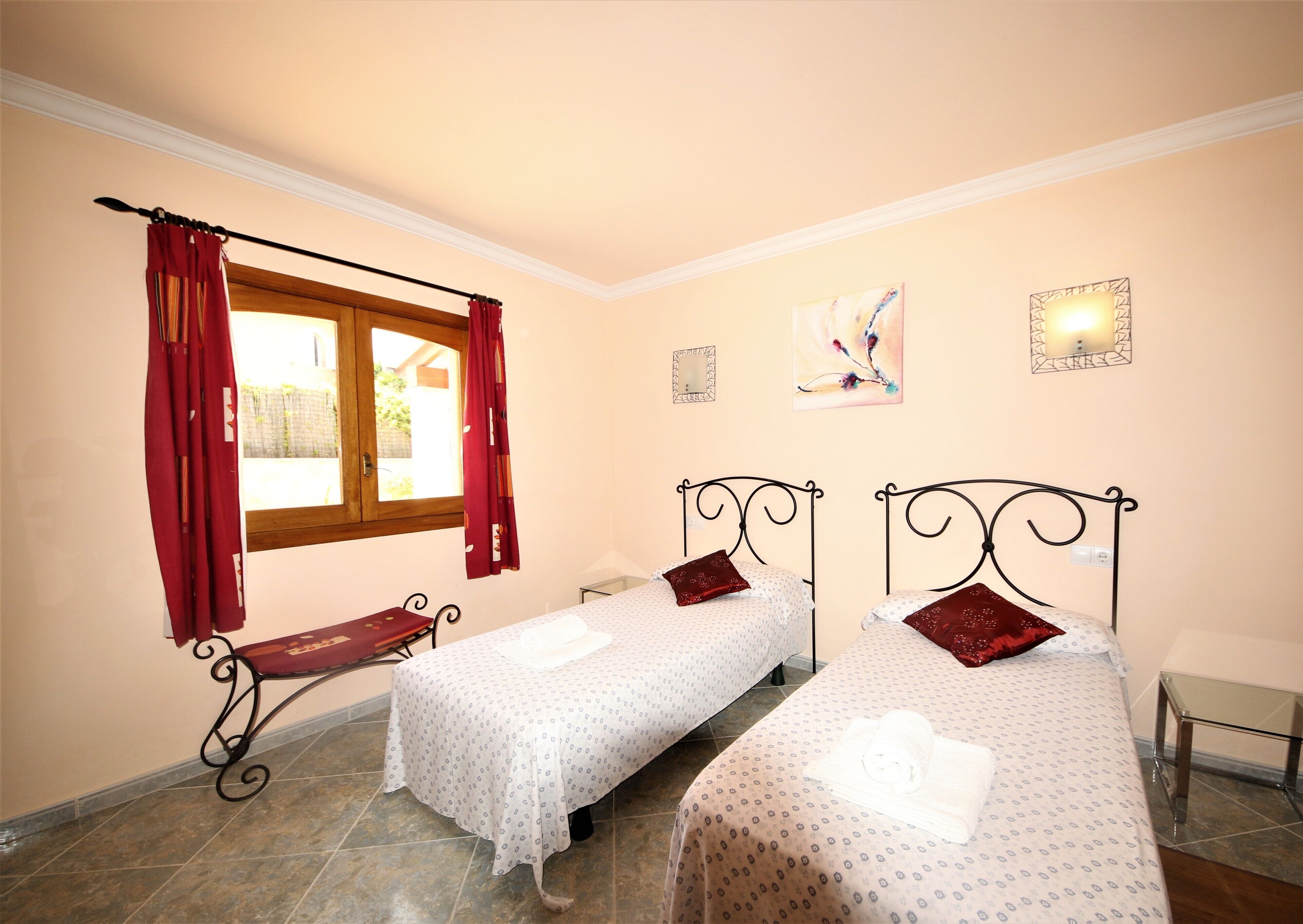 A bedroom with two single beds, a bench, a window with curtains, and artwork on the walls.