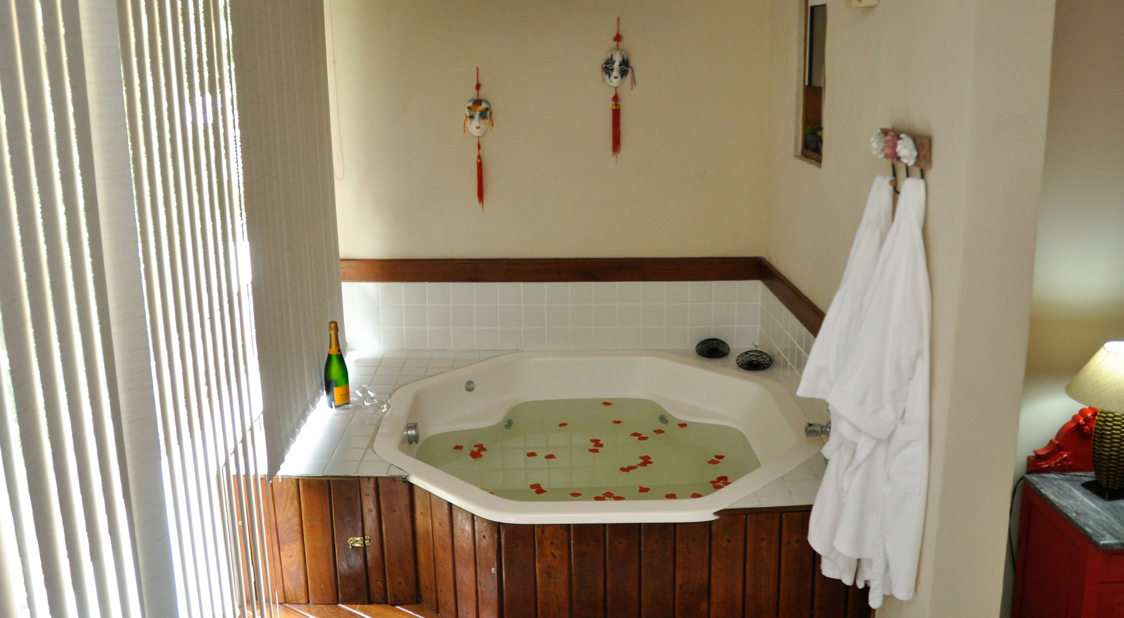 A bathroom with a wooden floor, a white bathtub with rose petals, a bottle of champagne, and a towel rack.