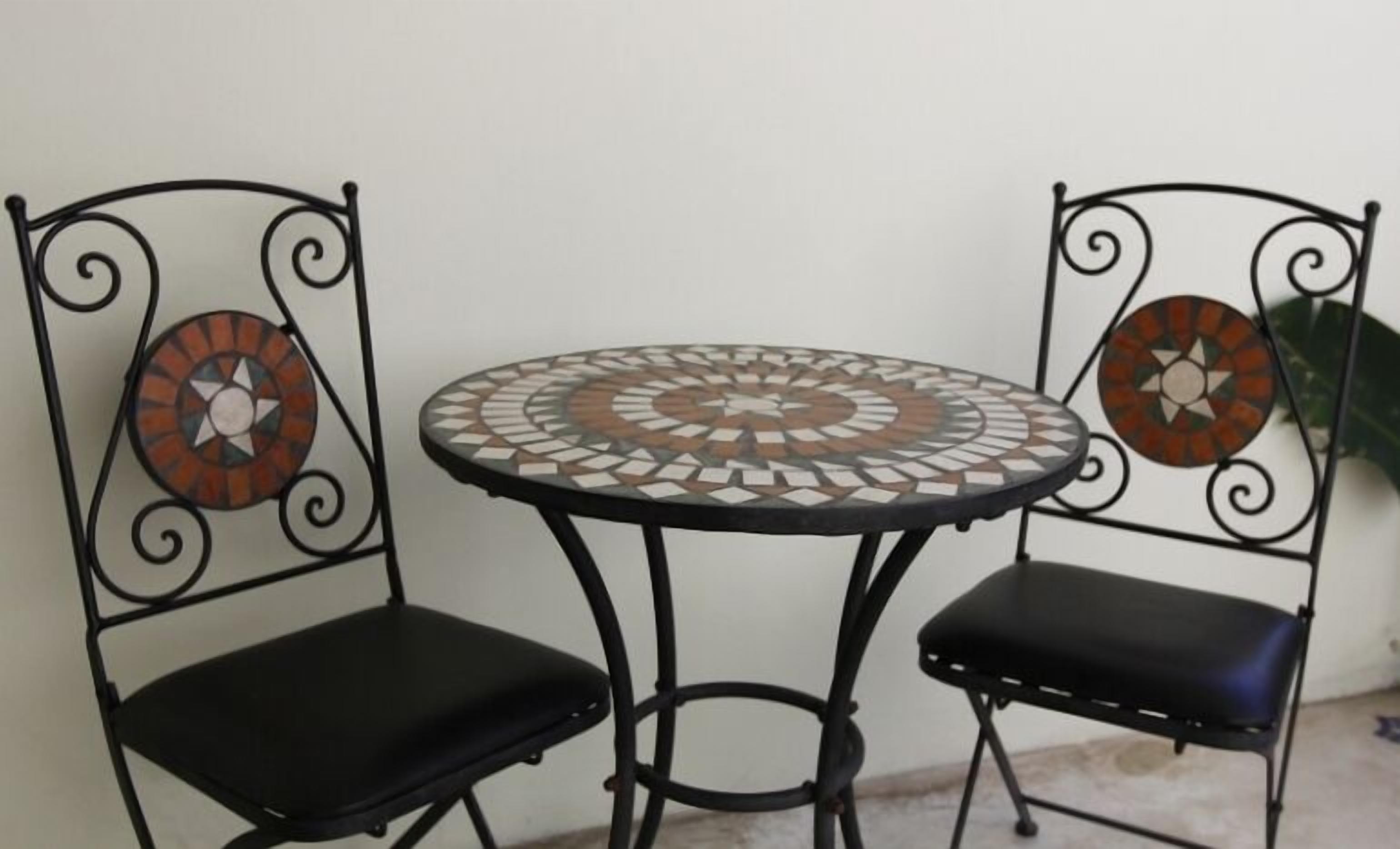 A small round table with mosaic tile design, flanked by two chairs with metal frames and cushioned seats.