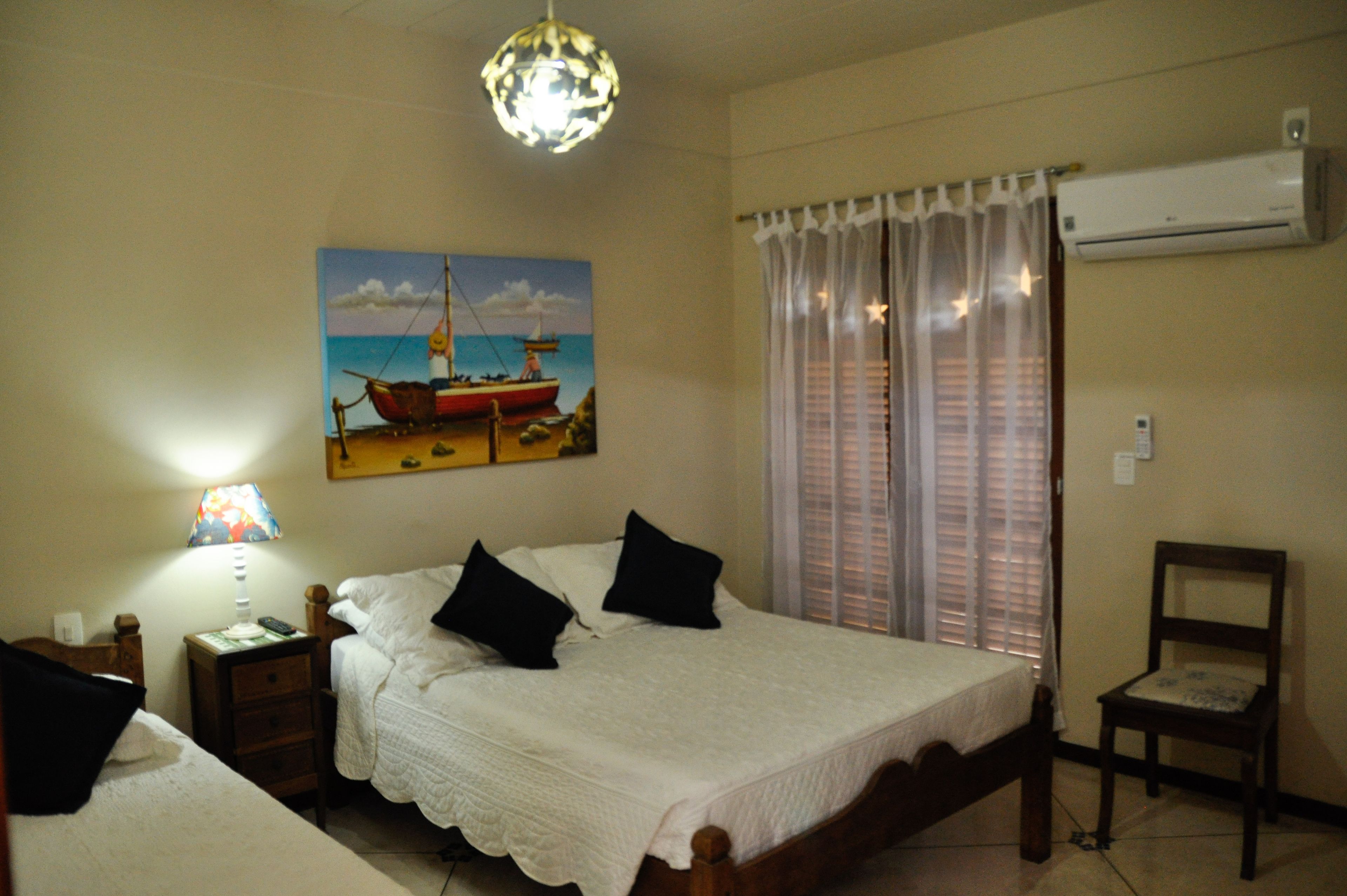 Un dormitorio con cama, mesita de noche, lámpara, silla, un cuadro de un barco en la pared y aire acondicionado.