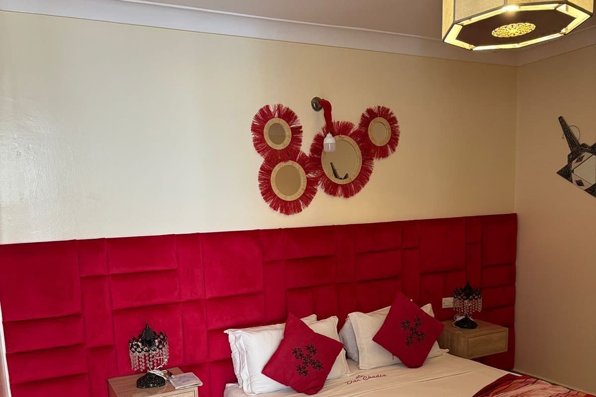 A bedroom with a red headboard, a bed with white bedding and red pillows, a nightstand with a lamp, and a wall-mounted light fixture.