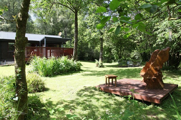 Une terrasse en bois avec une table et une grande sculpture d’arbre, le tout situé dans un jardin luxuriant et verdoyant, avec une cabane en arrière-plan.