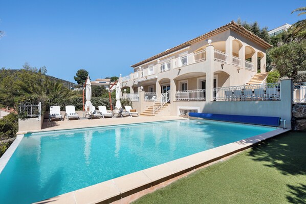 A large swimming pool with a white fence, a covered seating area with chairs, and a building with multiple balconies. - Villa 55 -Heated pool with exceptional view (Roquebrune-Sur-Argens)