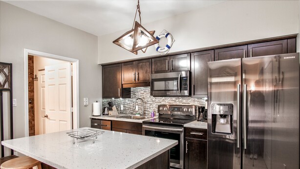 A modern kitchen with stainless steel appliances, a large island with stools, and a hanging light fixture. - Avalon Ground floor condo (clearwater)