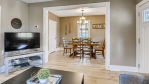 A modern living room with a flat-screen TV on a glass stand, a dining area with a wooden table and chairs, and a hallway leading to another room. - Charming Family & Corporate Retreat Near Broadmoor (Colorado Springs)
