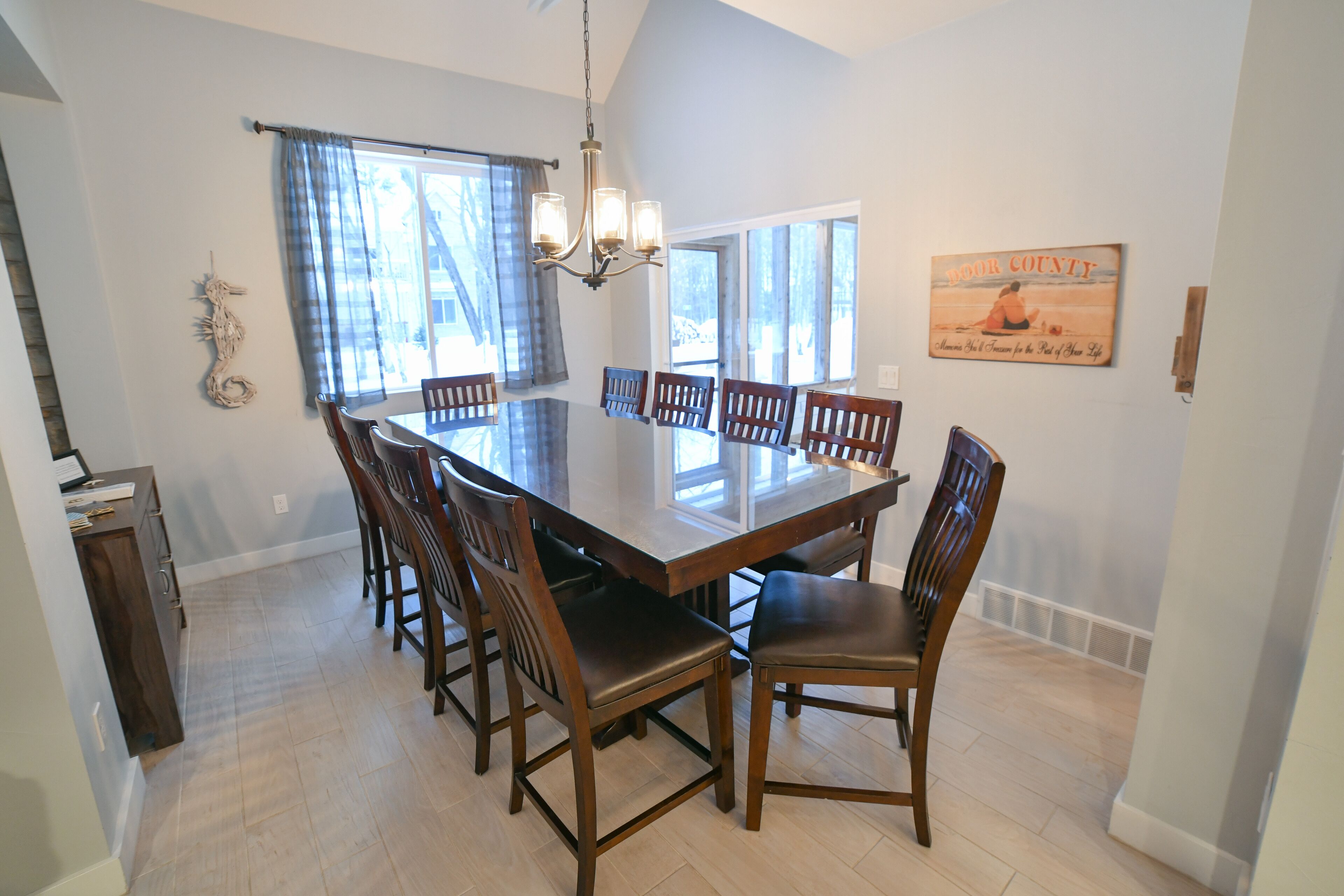 A dining area with a large rectangular table surrounded by wooden chairs, a chandelier, and large windows with curtains.