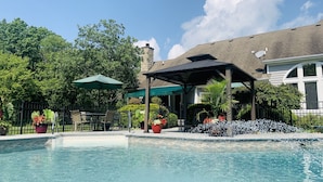Una piscina con un porche techado, rodeada de abundante vegetación y una chimenea de piedra.