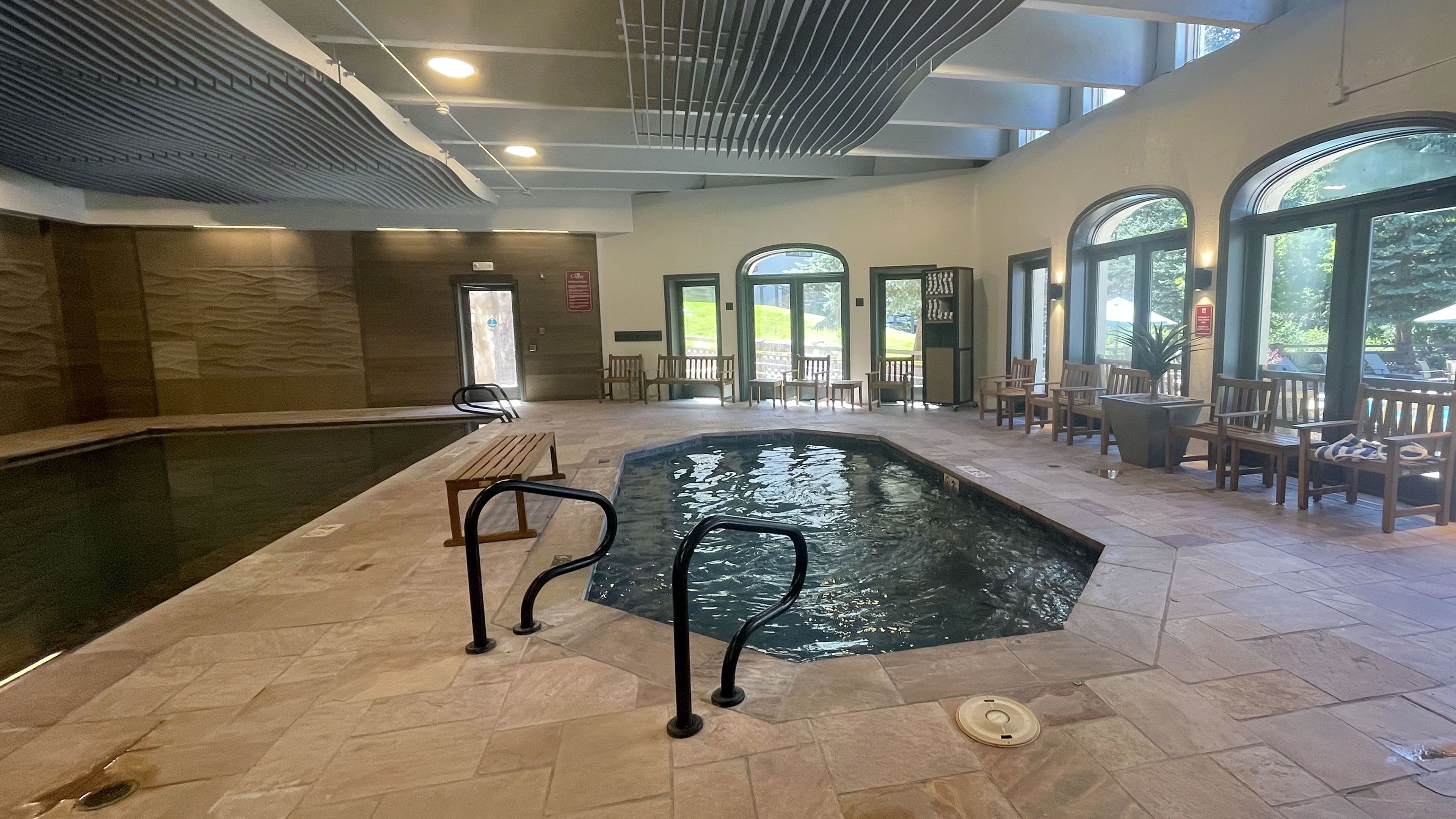 An indoor swimming pool with benches and chairs around it, large windows, and a ceiling with a unique design.
