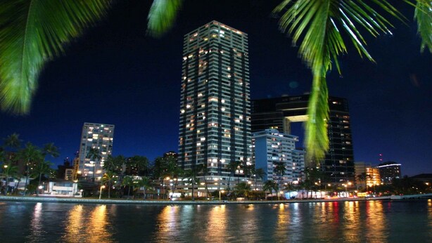 A nighttime cityscape with high-rise buildings along a waterfront, palm trees, and reflections on the water. - Renovated Specious Studio with Great View (Honolulu)