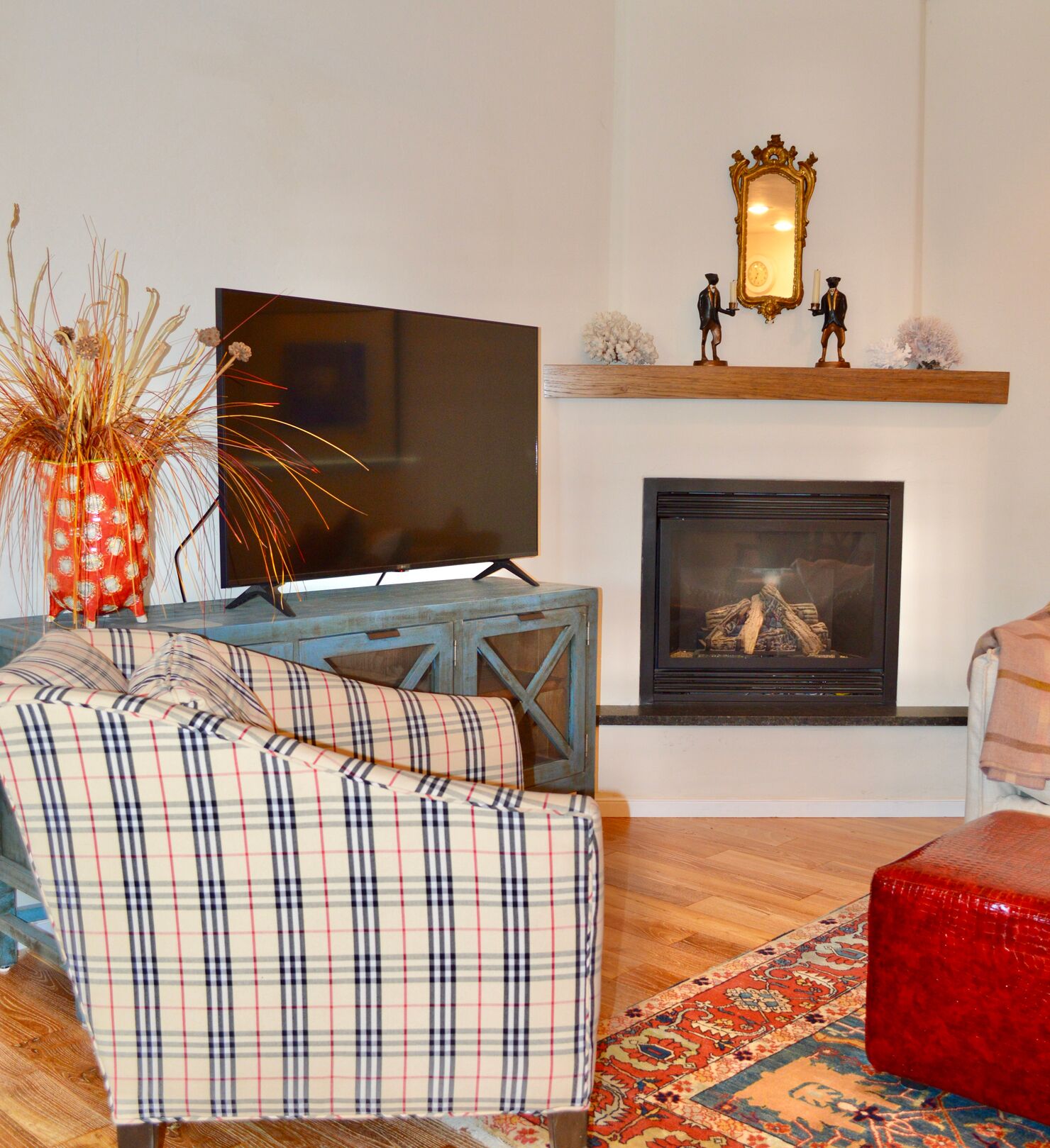 Un salon avec une télévision, une cheminée, un canapé à carreaux et un pouf rouge.