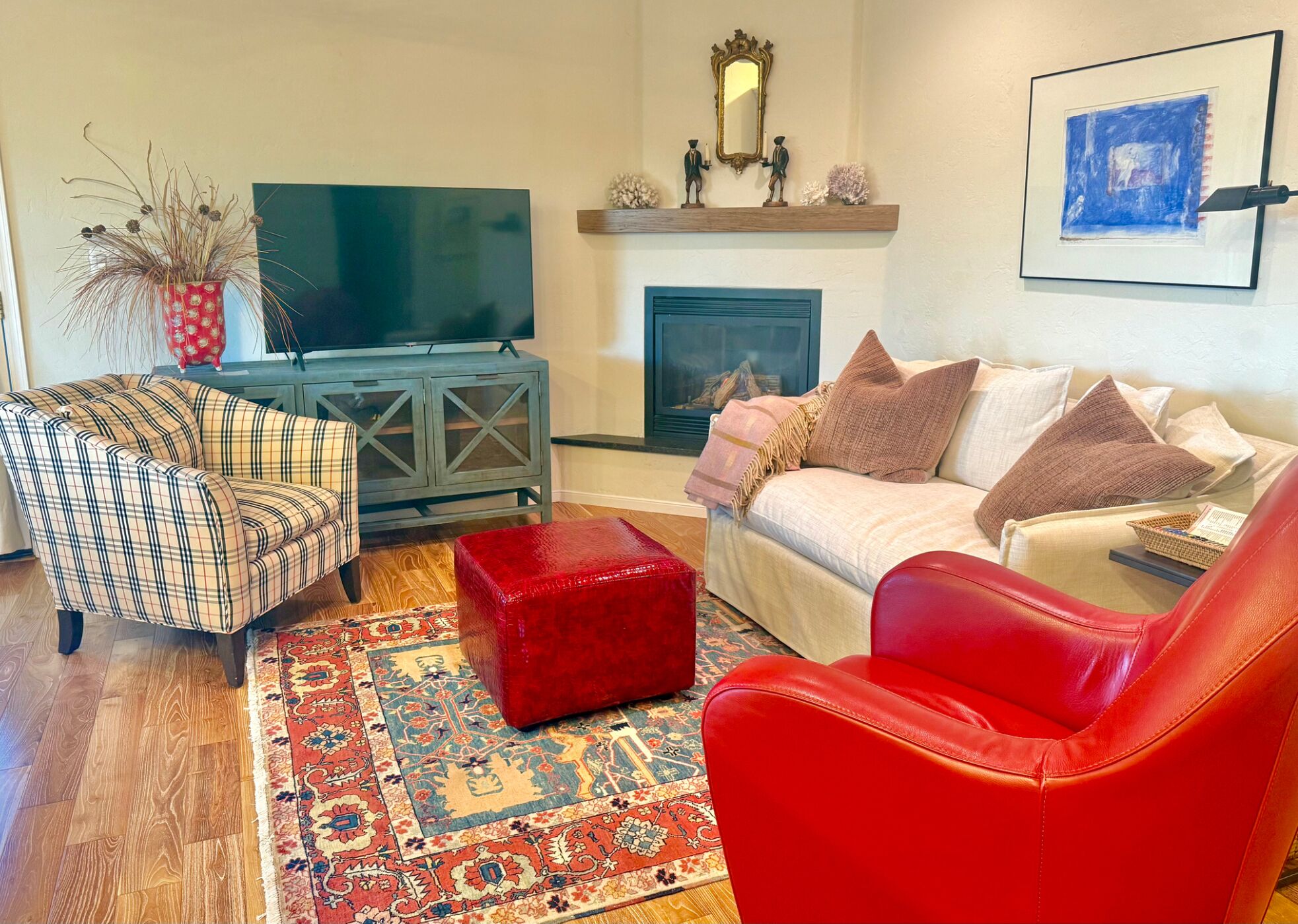 Un salon comprenant un fauteuil à carreaux, un pouf rouge, un fauteuil en cuir rouge, un canapé beige orné de coussins à motifs, un téléviseur à écran plat, une cheminée et une œuvre d’art encadrée accrochée au mur.