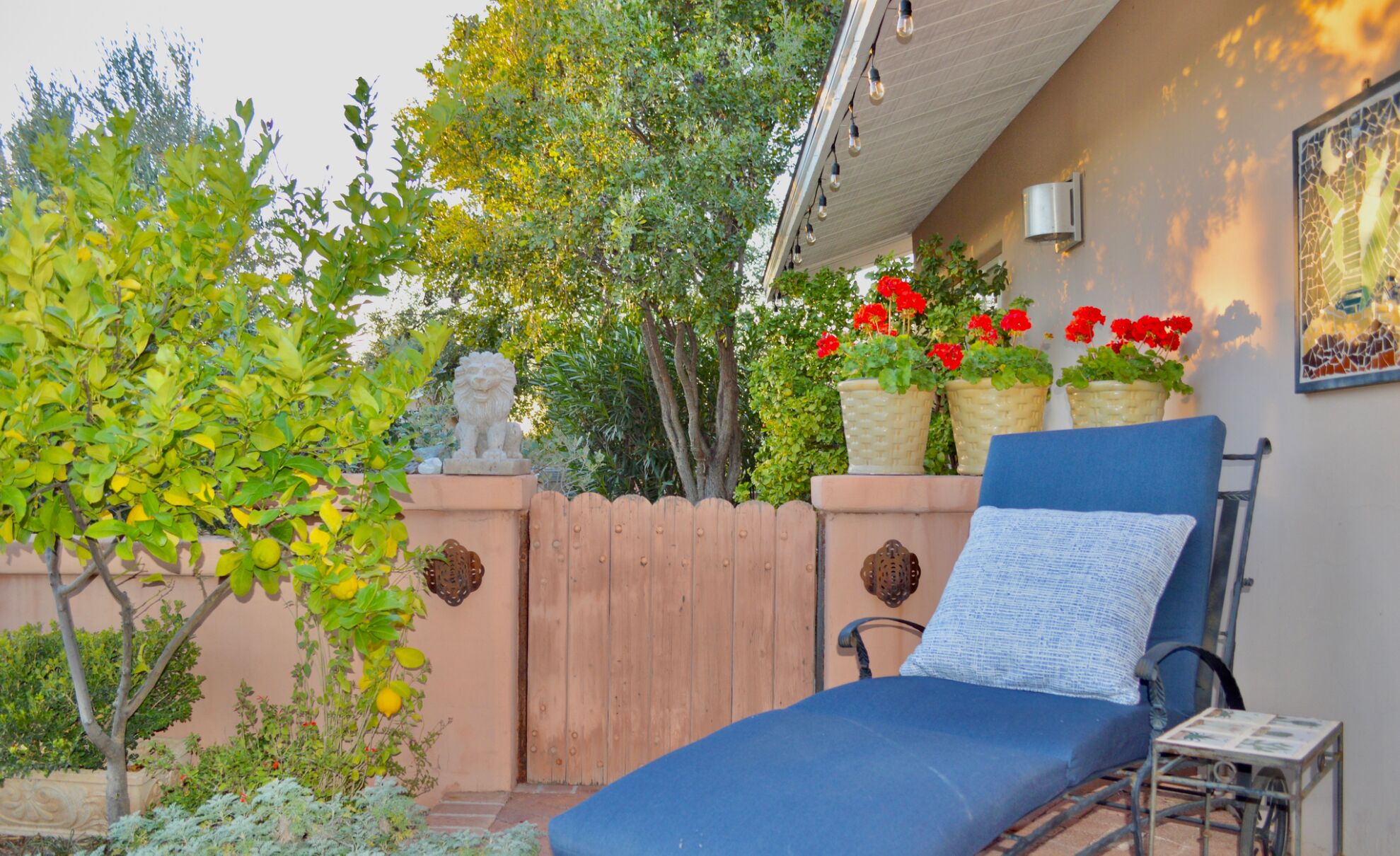 Un patio iluminado por el sol con una silla de lounge azul, una valla de madera y macetas con flores rojas.