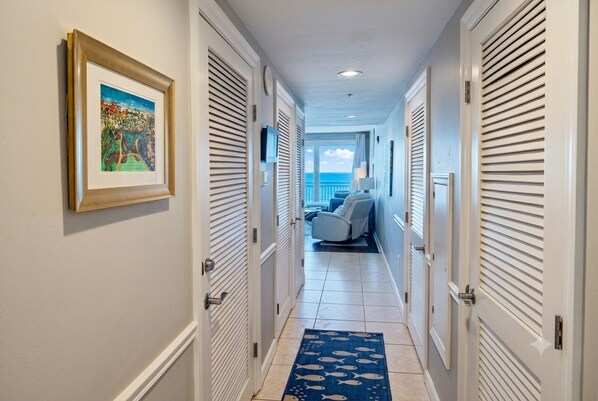 A hallway with closed doors, a framed picture on the wall, a blue rug with fish patterns, and a view of a balcony with a chair. - ☀️SUMMER ISN’T OVER! Escape to Sterling Shores in Destin 🌊🏖️ make one more memory! (Destin)