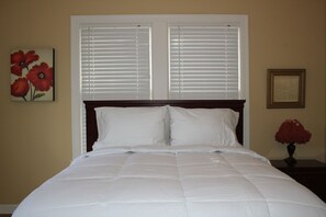 A neatly made bed with white bedding, a wooden headboard, and a window with blinds.