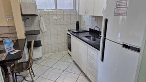 A compact kitchen with white cabinets, a black countertop, and a stainless steel sink. There's a small dining area with a table and chairs, and a trash bin is visible. - Apartamentos Adriático Guarujá (Guarujá)