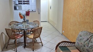 A dining area with a round glass table and wicker chairs, a kitchen with white cabinets and a microwave, and a hallway leading to a door. - Apartamentos Adriático Guarujá (Guarujá)