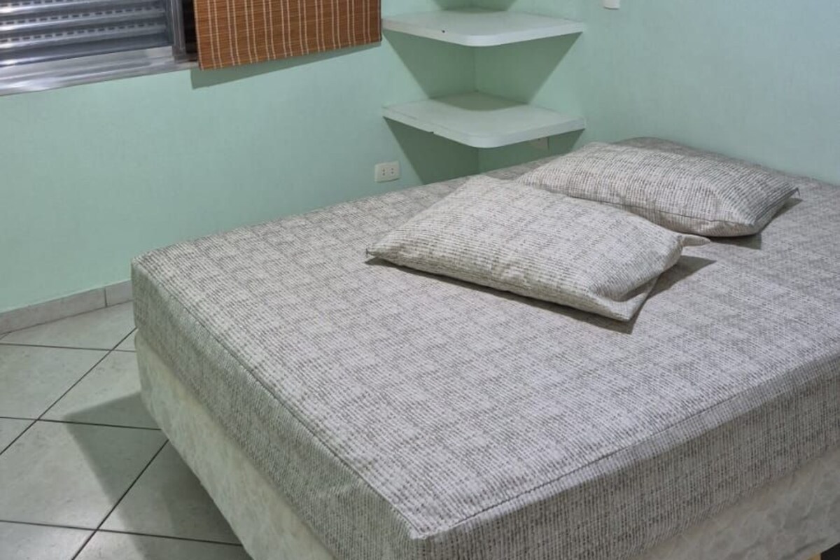 Une chambre à coucher comprenant un lit, un ventilateur de plafond, une fenêtre avec des stores et une étagère fixée au mur.