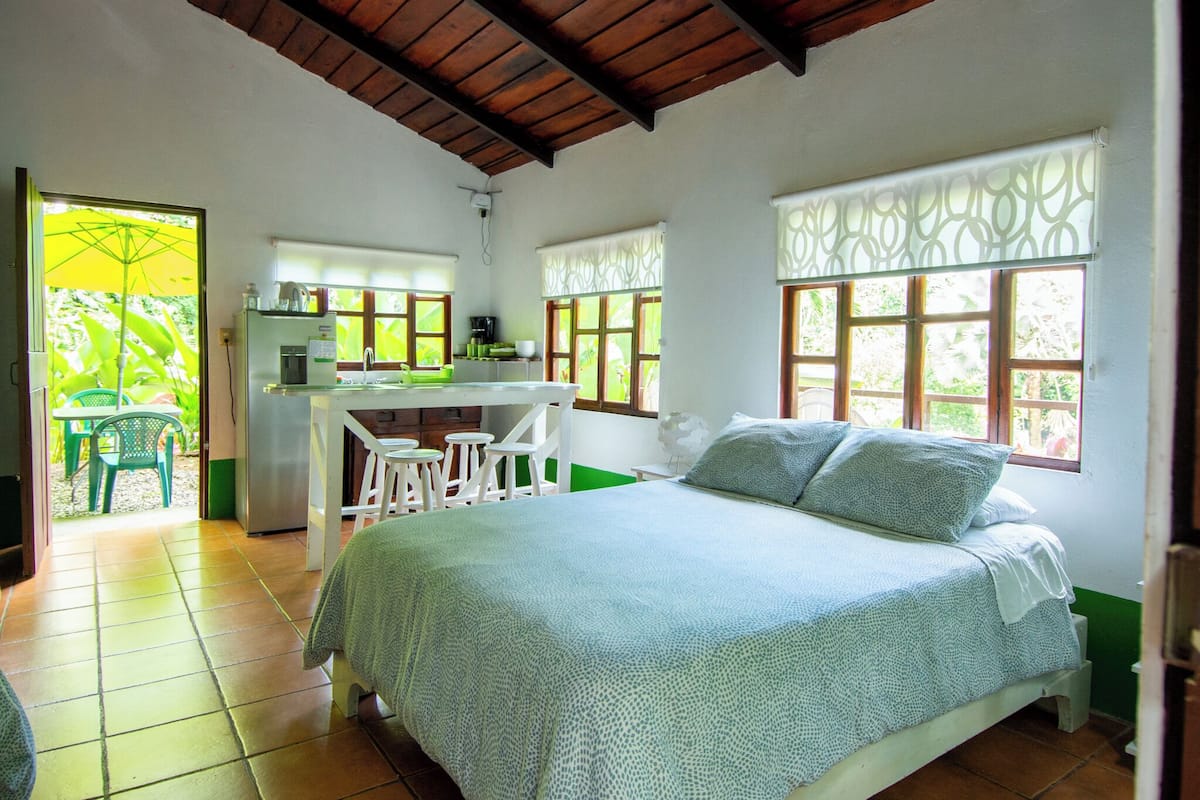 A bedroom with a bed, a desk, and a chair. There is a kitchen area with a refrigerator and a sink.