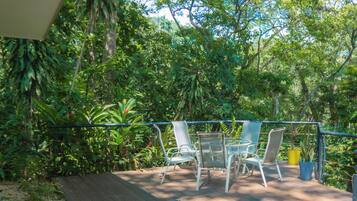 An outdoor deck with a wooden floor, surrounded by lush greenery and trees. There are two glass-top tables with metal chairs.