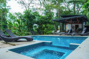 Piscina con sillas de descanso y un área de estar cubierta, rodeada de exuberante vegetación.
