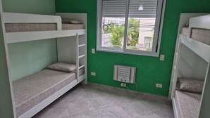 A room with bunk beds, a window with blinds, and a wall-mounted air conditioning unit. - Apartamento Baia Blanca Guarujá (Guarujá)