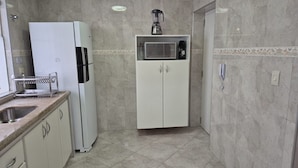 A compact kitchen with a refrigerator, microwave, and a coffee maker on a shelf. - Apartamento Baia Blanca Guarujá (Guarujá)