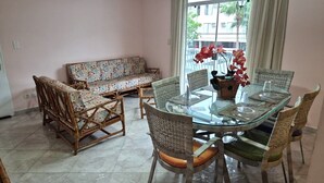A dining area with a glass-top table, wicker chairs, and a floral sofa. - Apartamento Baia Blanca Guarujá (Guarujá)