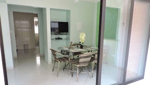A dining area with a glass table, wicker chairs, and a television in the background. - Apartamento Lester Primo Guarujá (Guarujá)