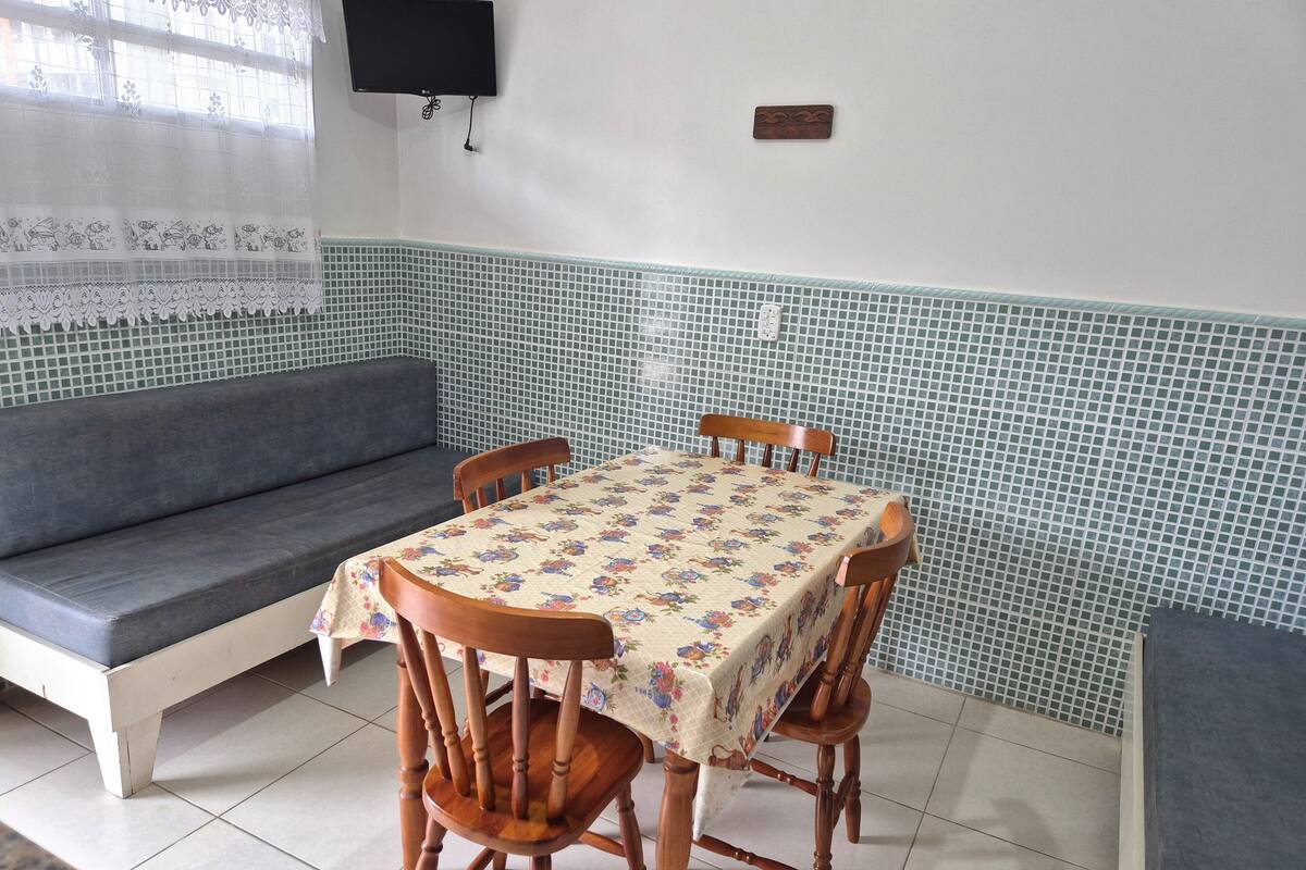 A dining area with a table covered in a floral tablecloth, surrounded by wooden chairs, a grey sofa, and a tiled backsplash.