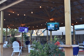 An outdoor patio area with wooden chairs and tables, a covered structure with string lights, and a digital screen displaying information. - The Oak Motel (La Grange)