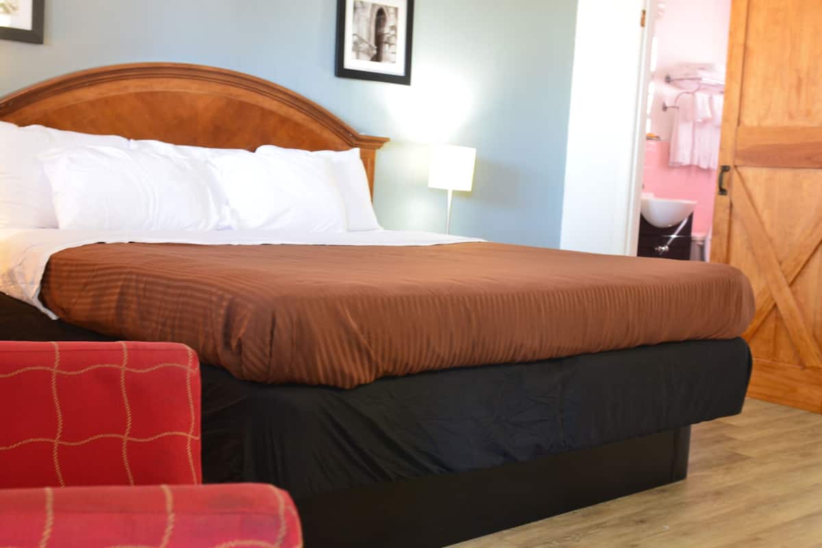 Una cama bien hecha con cabecera de madera, una silla roja y una mesita de noche con lámpara.