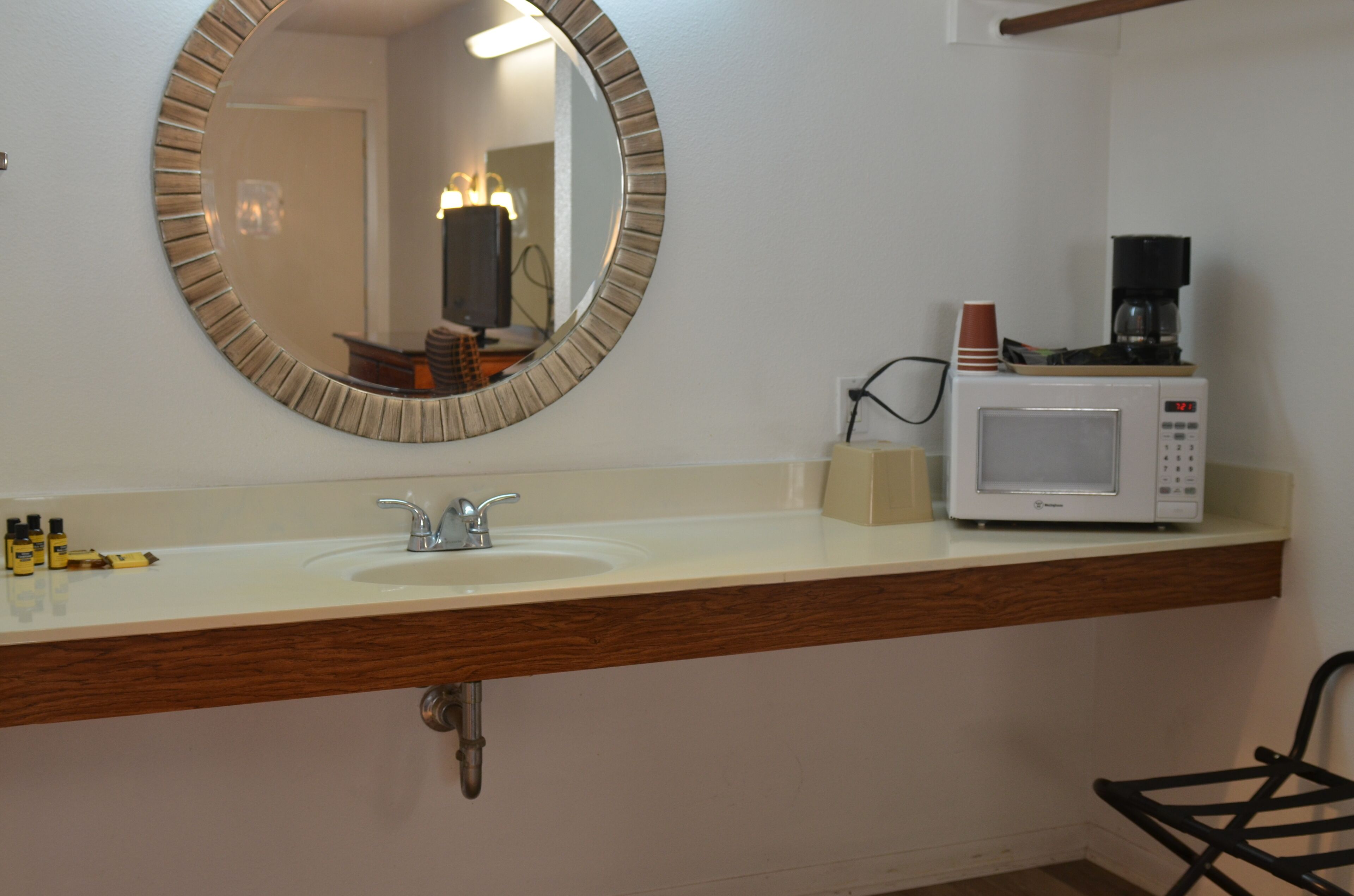 A bathroom with a round mirror, a sink, a coffee maker, and toiletries on the counter.