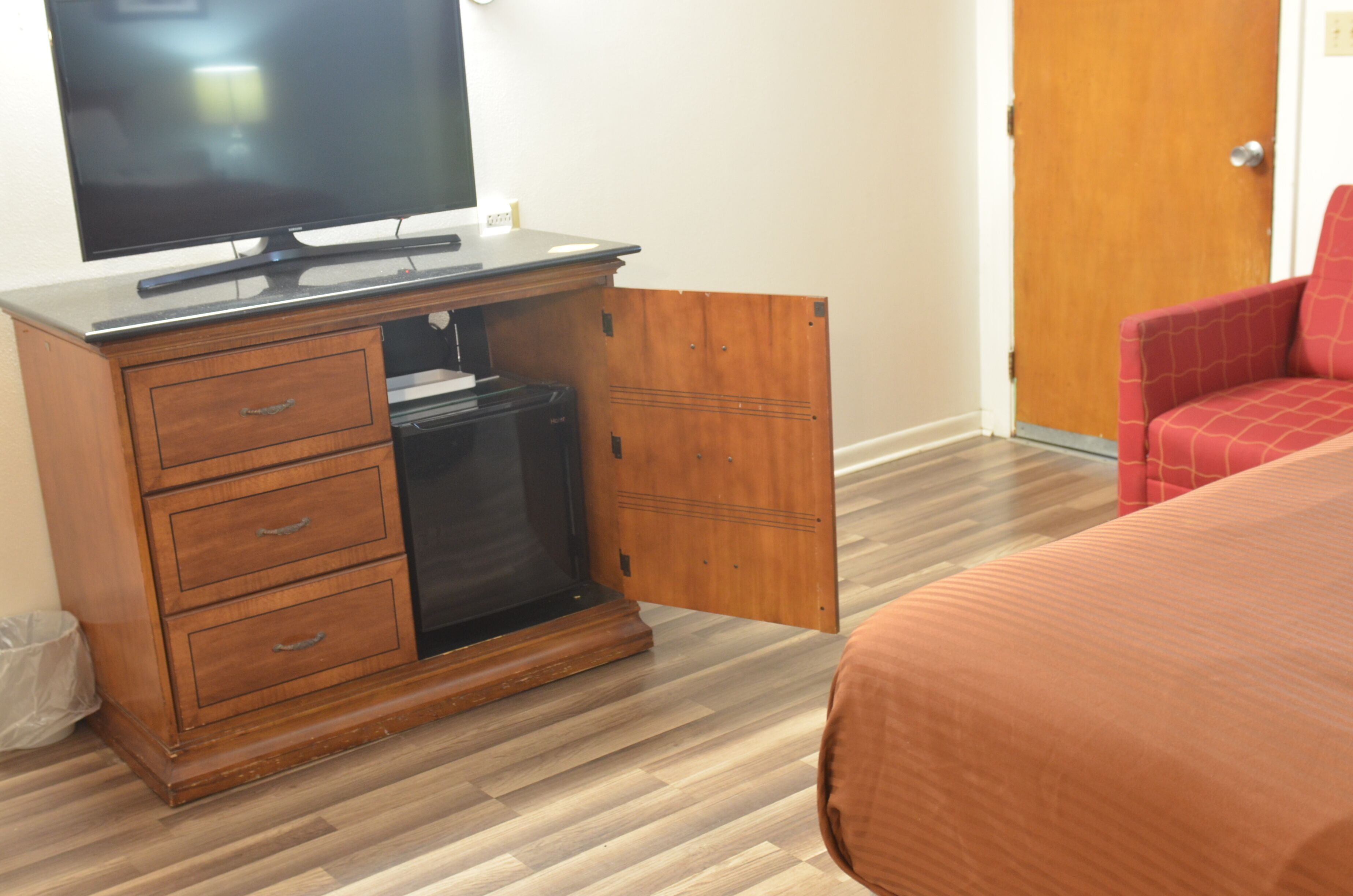 A hotel room with a wooden cabinet, a television, a red chair, and a bed with a brown bedspread.