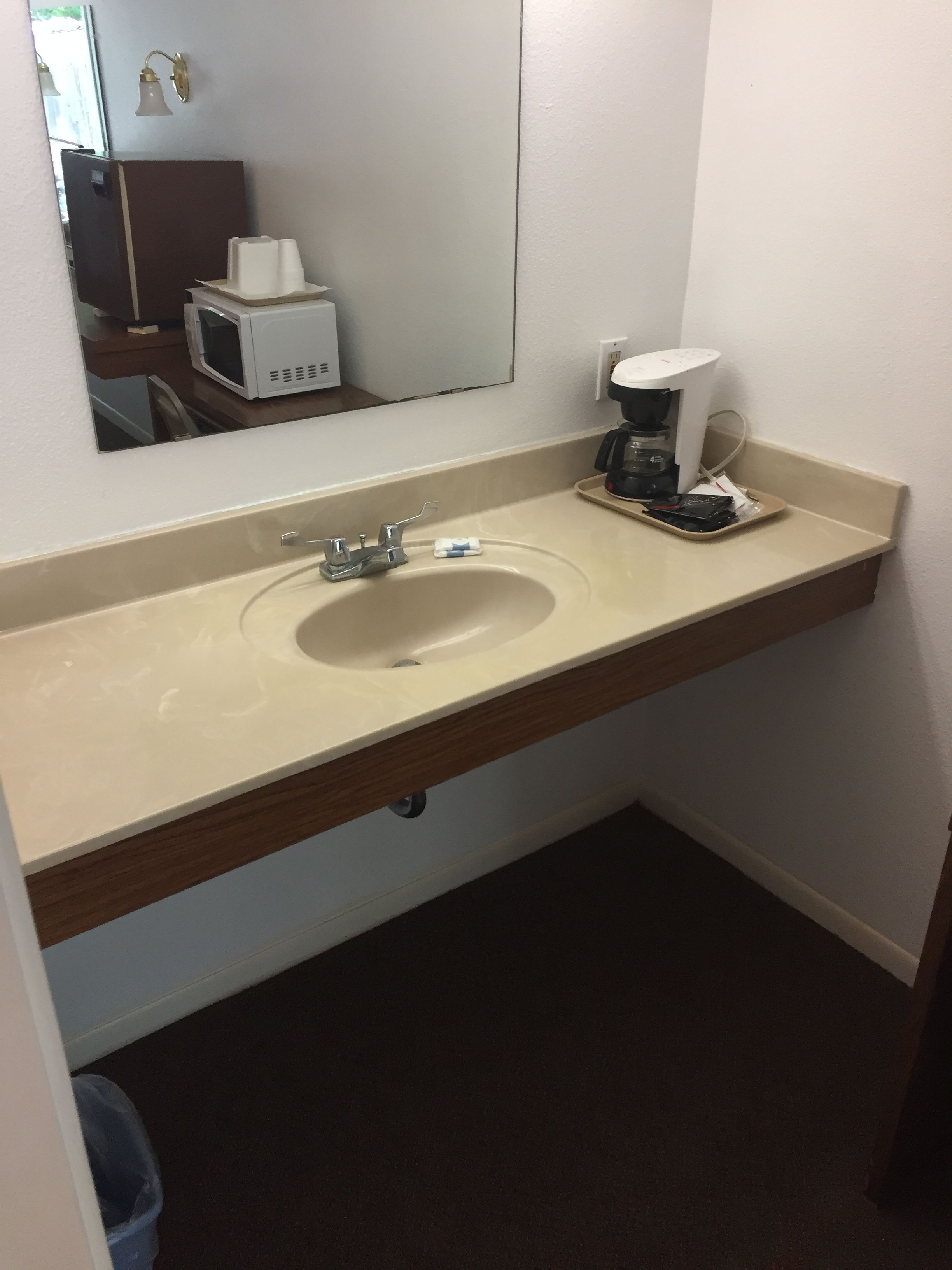 Un baño con lavamanos color beige, un espejo, una cafetera y un cubo de basura.