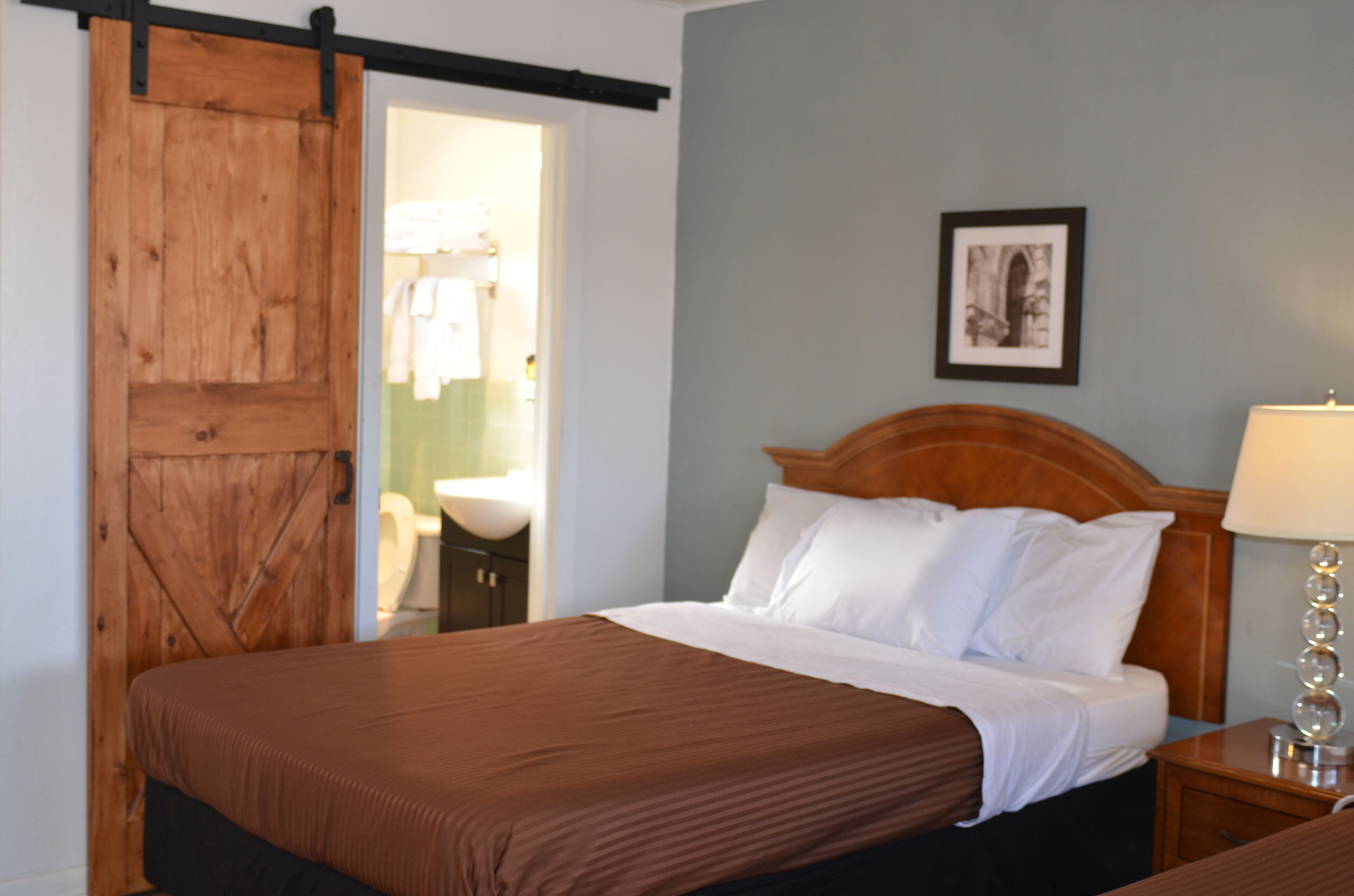 A hotel room with a wooden bed, a nightstand with a lamp, a framed picture on the wall, and a sliding door leading to a bathroom.