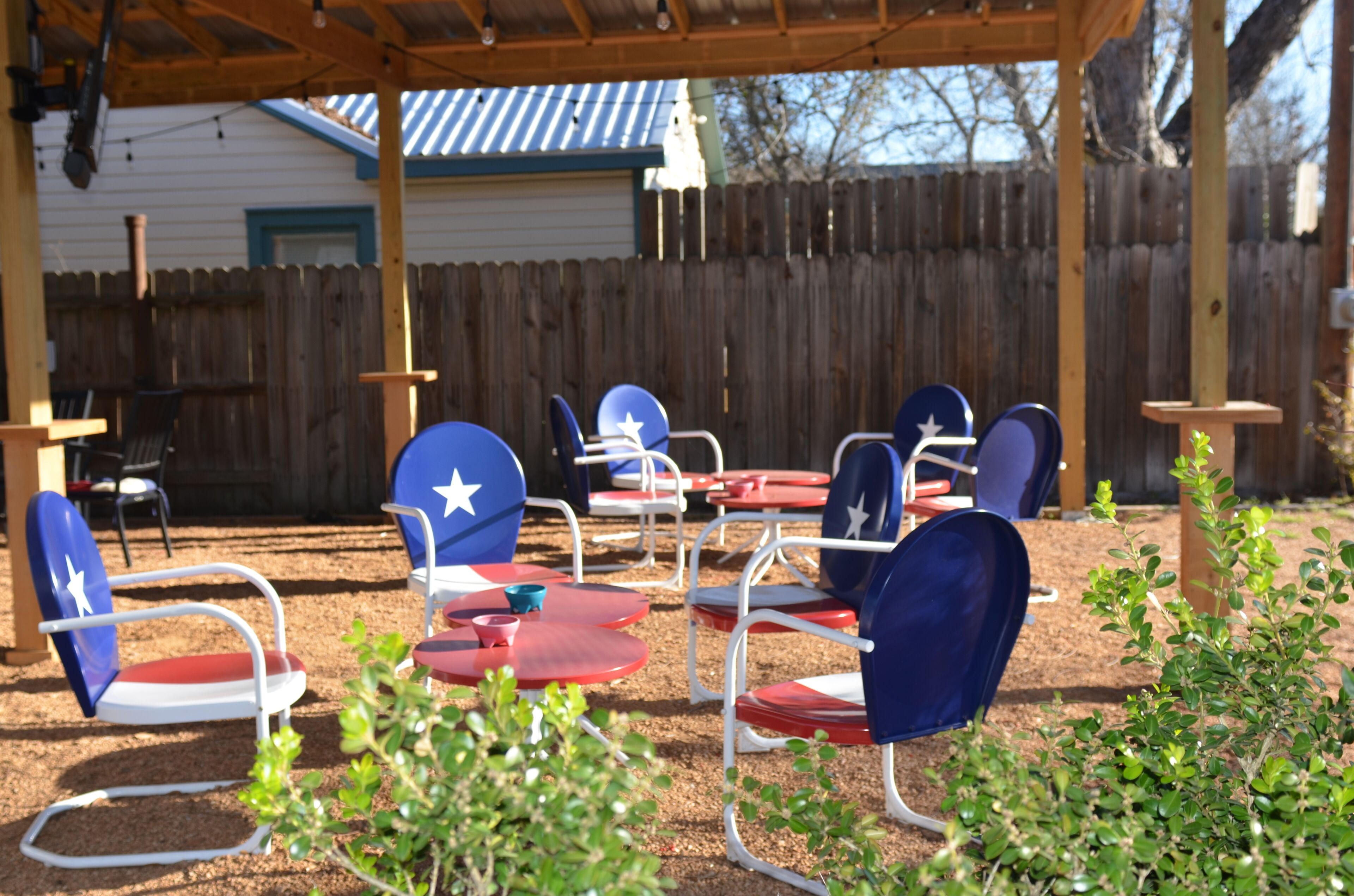 A covered outdoor seating area with metal chairs and tables, a wooden fence in the background, and a building with a blue roof.