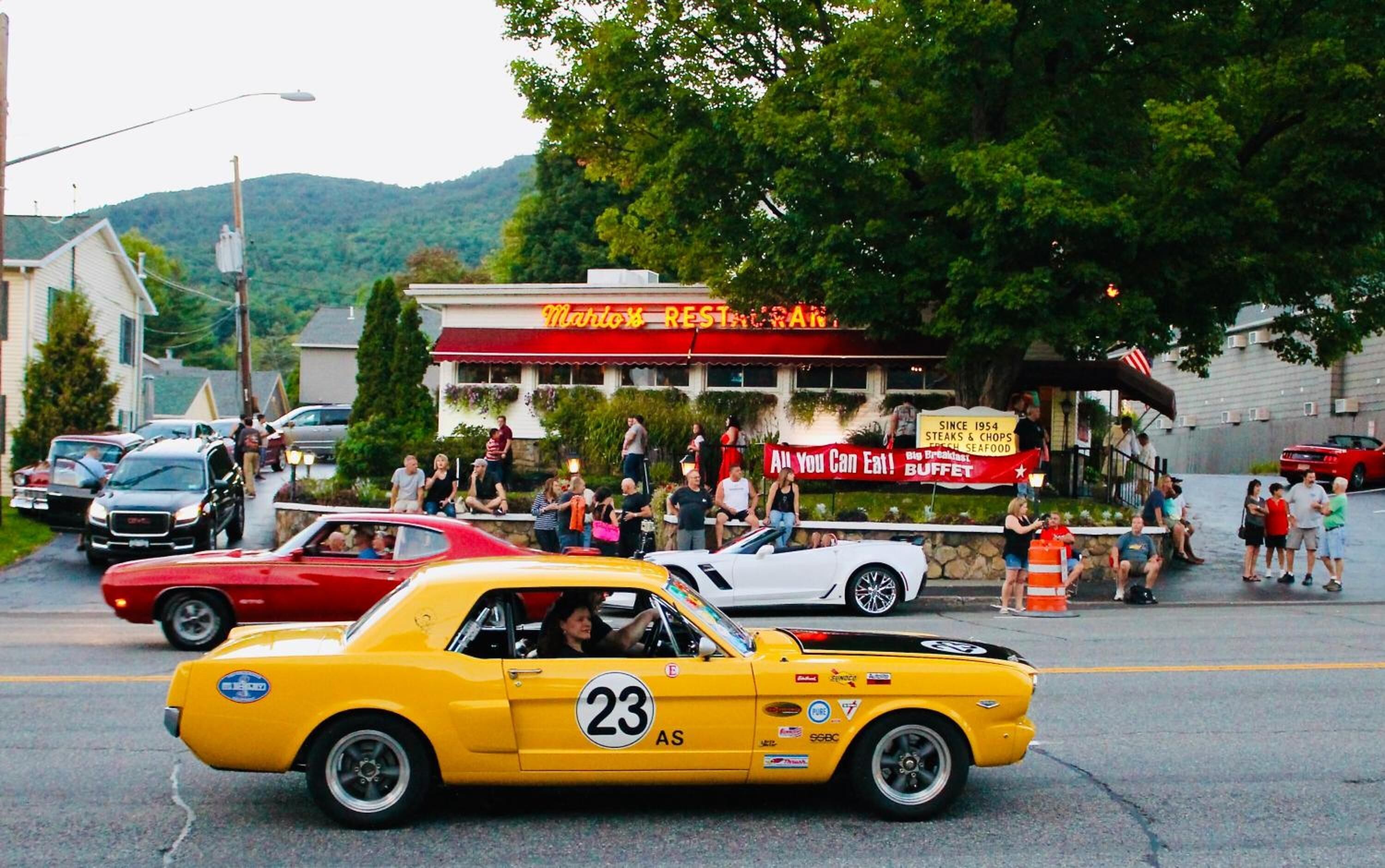 Un coche de carreras antiguo color amarillo con el número 23 en la carrocería, estacionado en una calle con gente y un restaurante al fondo.