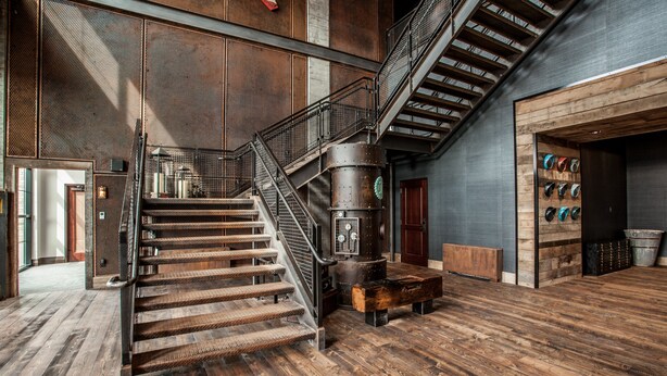 A spacious lobby with a wooden staircase, a large industrial-style heater, and a wall displaying various hats. - Ironworks Hotel Indy (Indianapolis)