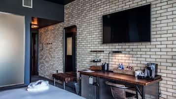 A modern hotel room with a brick wall, a flat-screen TV, a wooden bar area with a coffee machine, and a large bed.