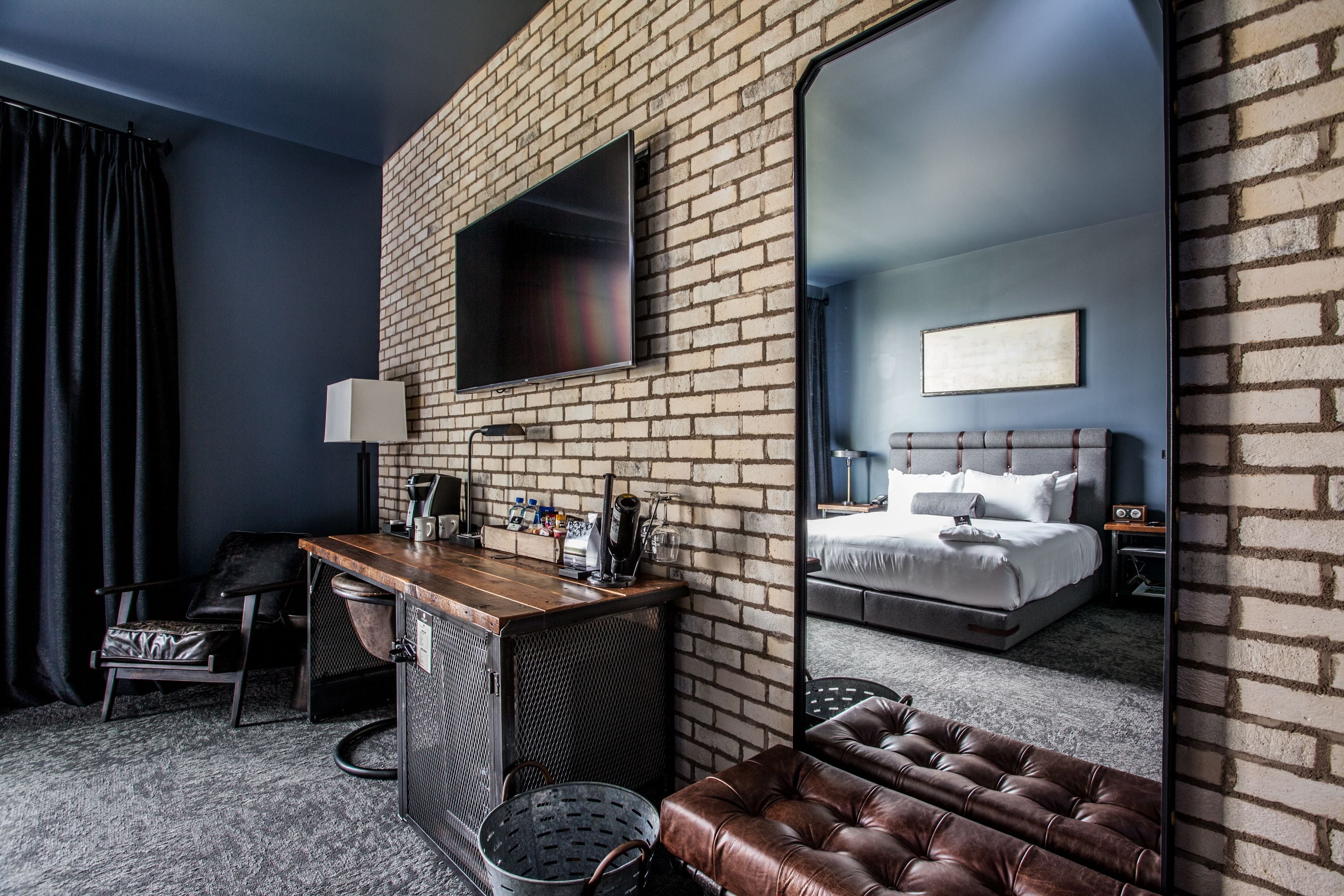 A modern hotel room with a brick wall, a large mirror, a desk with a lamp, a leather sofa, and a bed with a headboard.