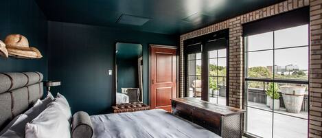 A modern hotel room with a large bed, a wooden bench, a mirror, and a view of the city through large windows.