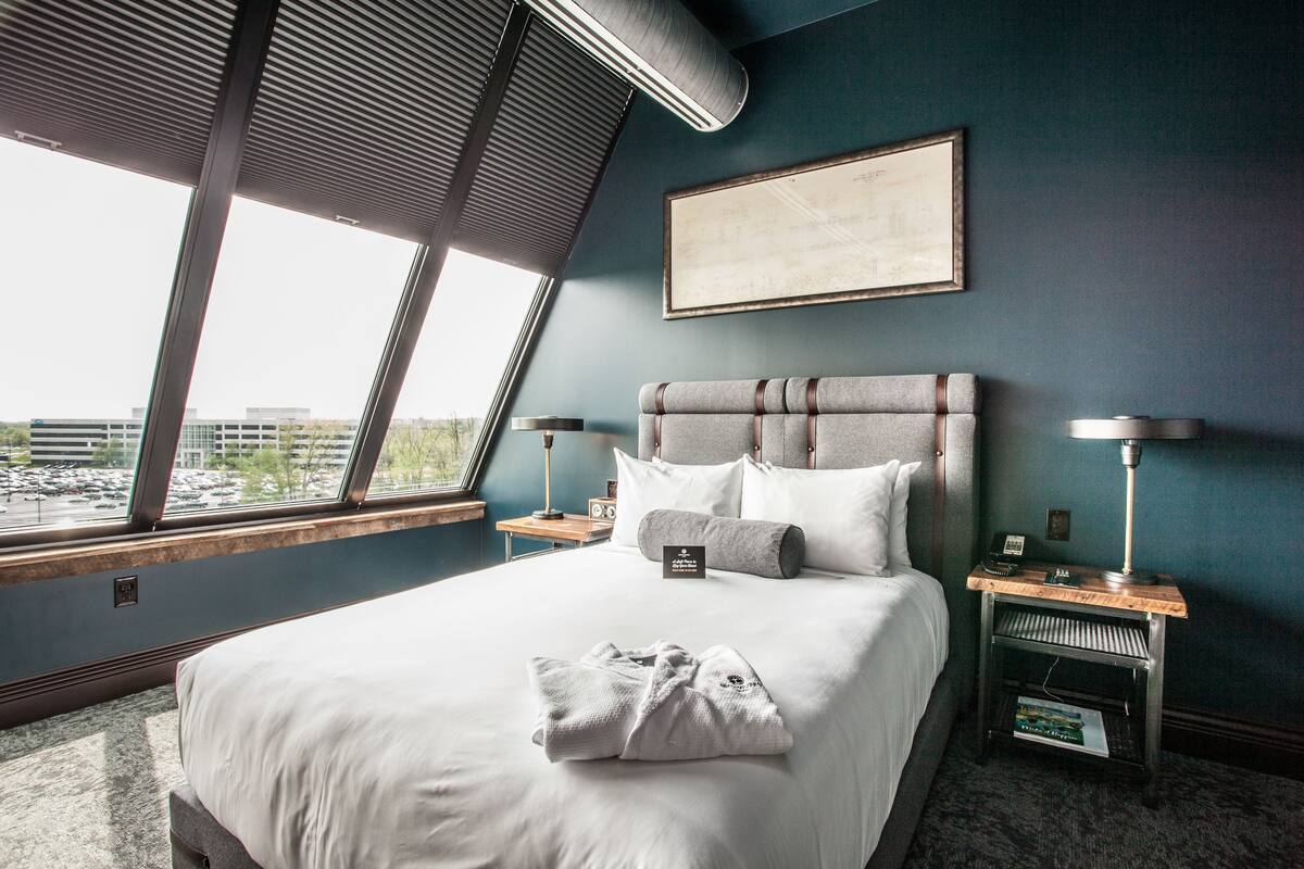 Ein Hotelzimmer mit einem großen Bett, Blick durch ein Fenster, einem gerahmten Bild an der Wand und einem Nachttisch mit Lampe.