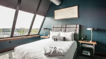 Habitación de hotel con una cama grande, vista a través de una ventana, un cuadro enmarcado en la pared y una mesita de noche con lámpara.