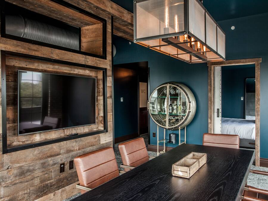 A modern room with a large dark wood table, leather chairs, a large flat-screen TV mounted on a wooden wall, a circular mirror with a glass display, and a bed with a white headboard.