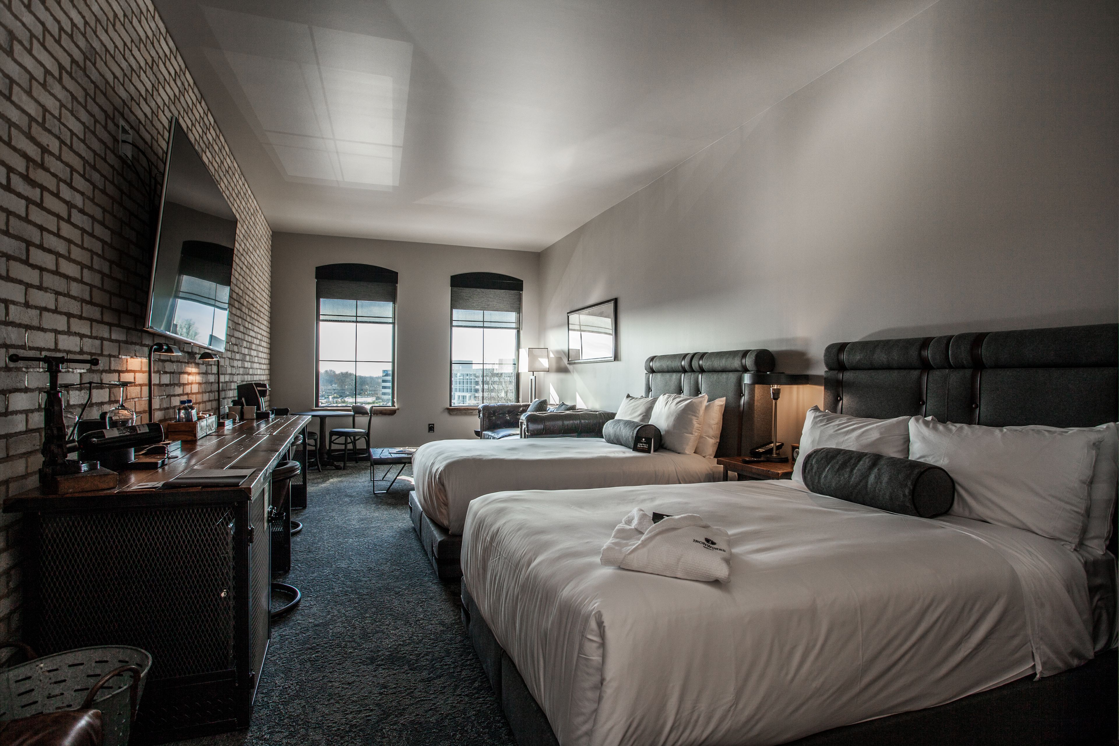 Quarto de hotel com duas camas, uma escrivaninha, parede de tijolos e janelas amplas.