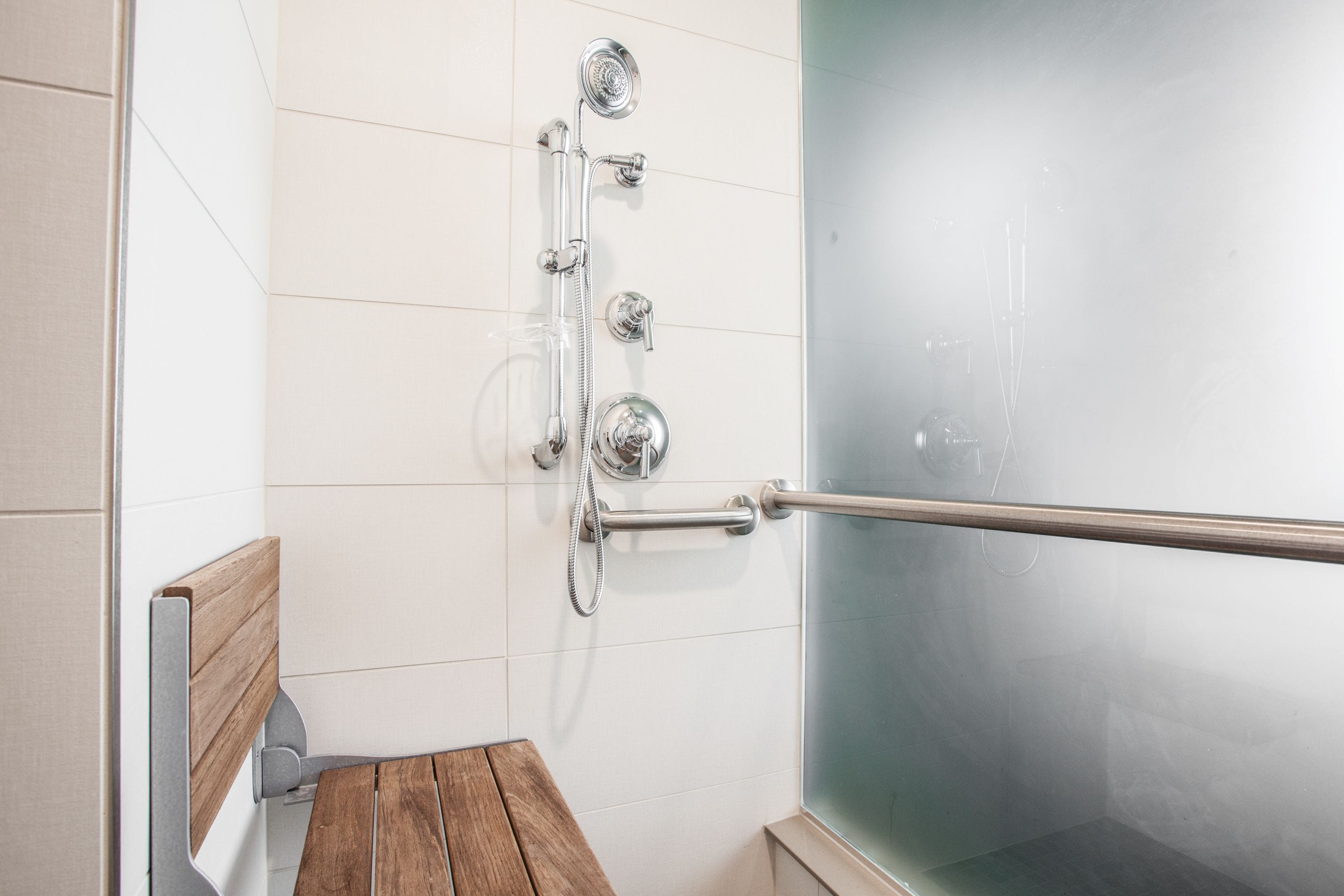 Un espace douche doté d’une porte en verre, d’un banc en bois et d’une barre d’appui.