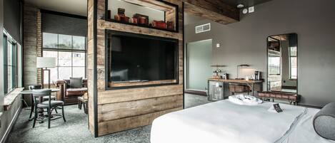 A modern hotel room with a large bed, a TV, a desk with a chair, and a seating area with a couch and chairs.